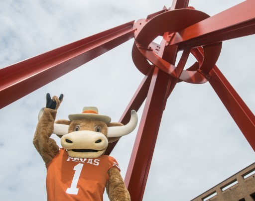 "Hook Em" UT's Longhorn Mascot, holds up a "hook em" gesture in front of Mark di Suvero's "Clock Knot" a large red metal sculpture that seems to meet in a mass in the upper right corner of the image