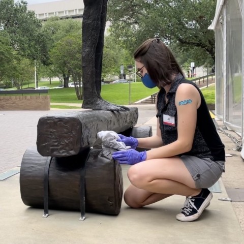 Ali Wysopal wearing a mask kneels down and rubs a bronze sculpture with a cloth.