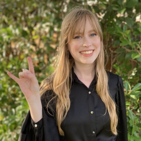 Caitlin Jones, a white woman with blonde hair smiles at the camera and holds up a "hook em" gesture