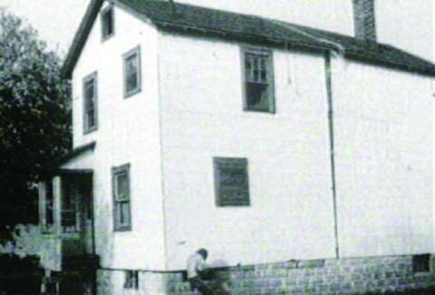 A rural two story house with a figure crouching in front