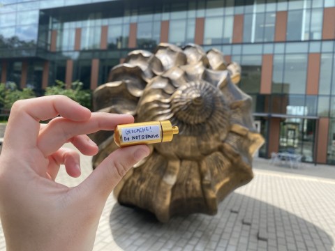 A hand holds up a small gold container that reads "Geocache! Do not remove" which is in front of Marc Quinn's "Spiral of the Galaxy" a large shell 