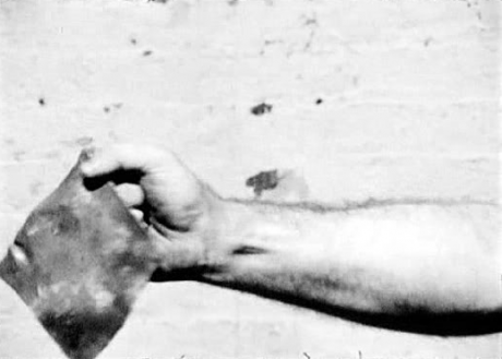 Film still of hand holding a piece of sheet metal