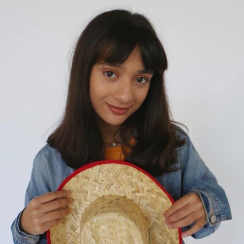 A headshot of Vega Shah. She has black hair and wears a denim shirt while holding a cowboy hat in the foreground of the photo